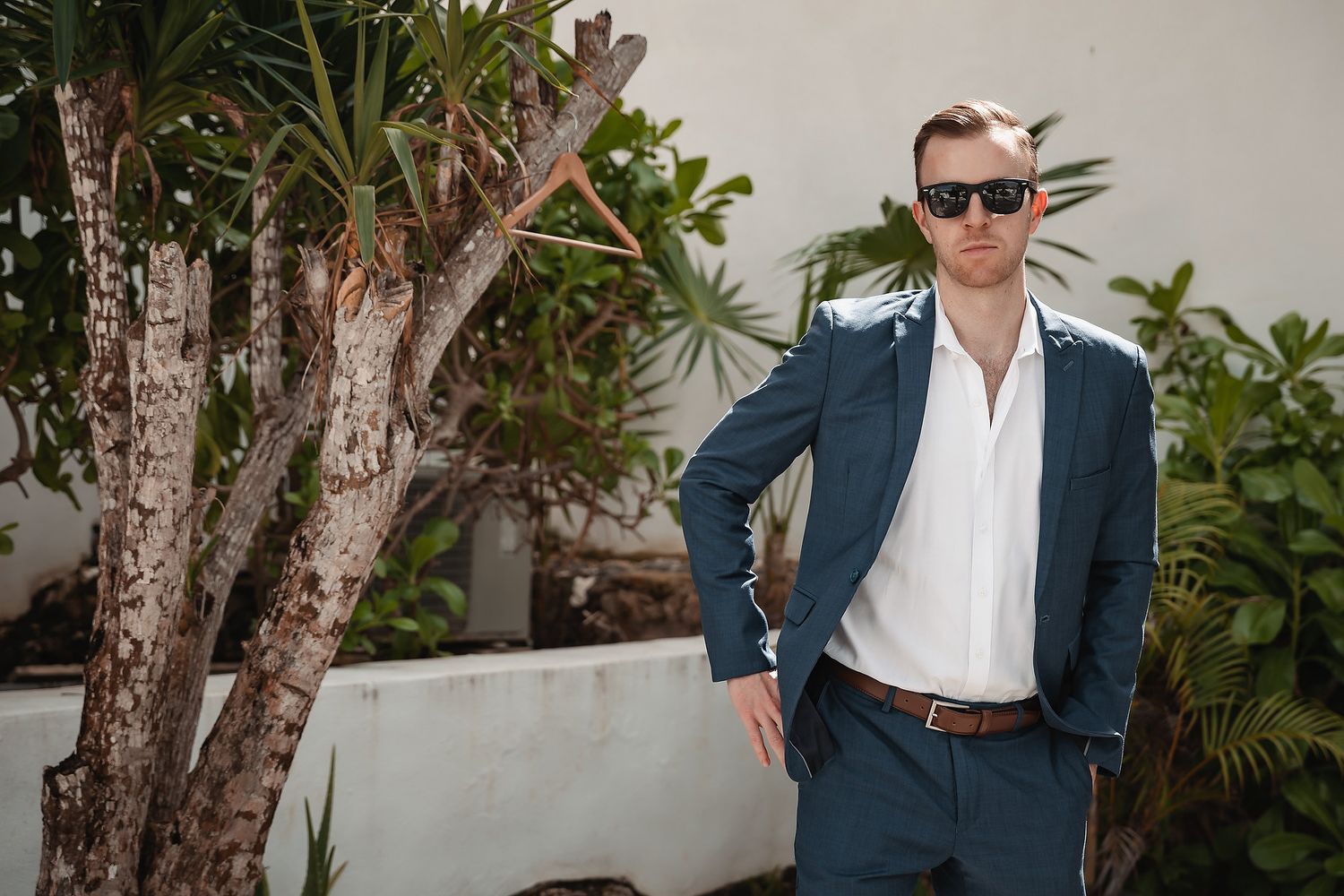 A stylish man in a navy suit and white shirt wearing sunglasses poses outdoors near tropical plants.