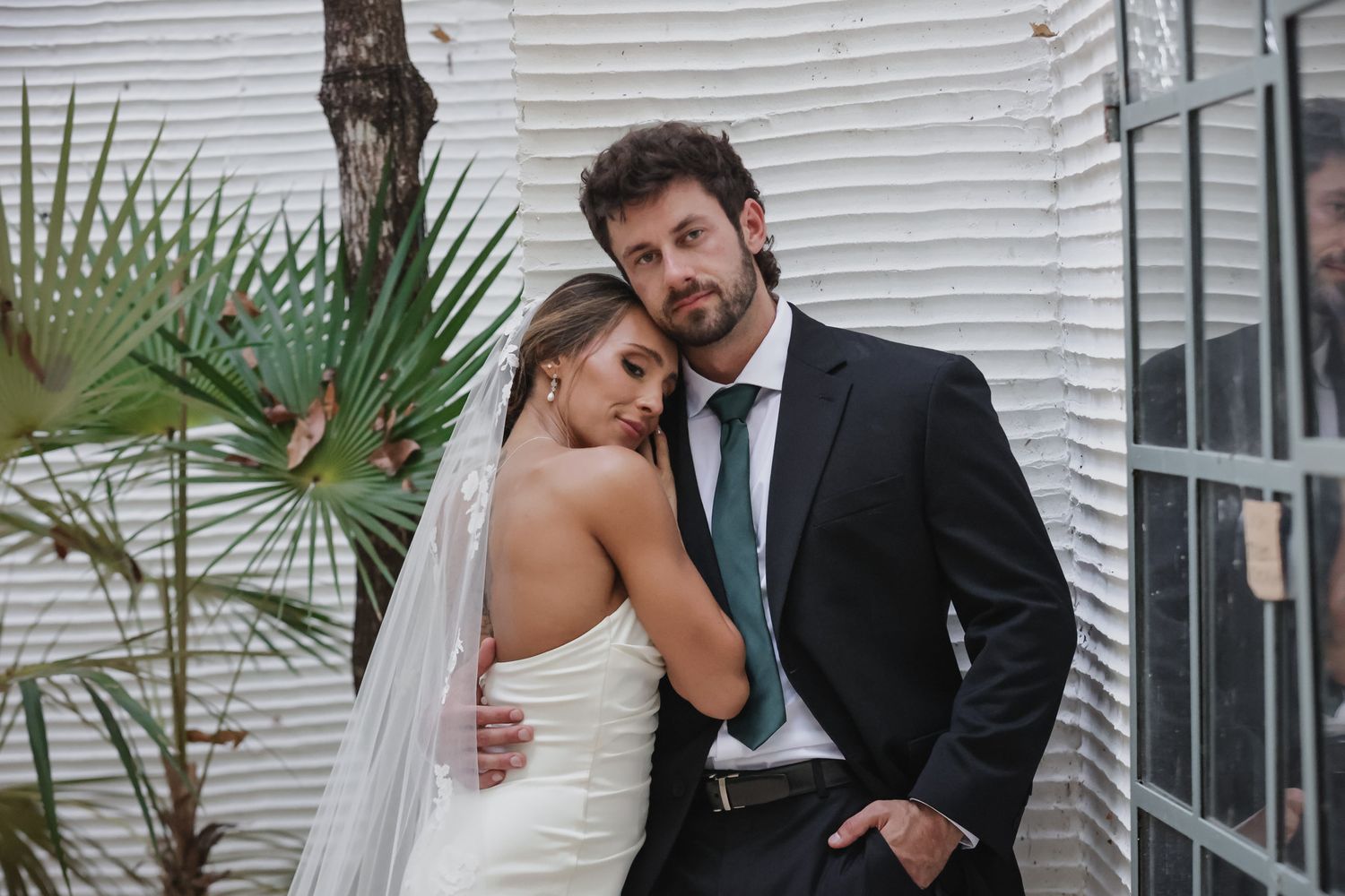 Elegant bride and groom posing outdoors, bride in white gown with veil, groom in dark suit with green tie.