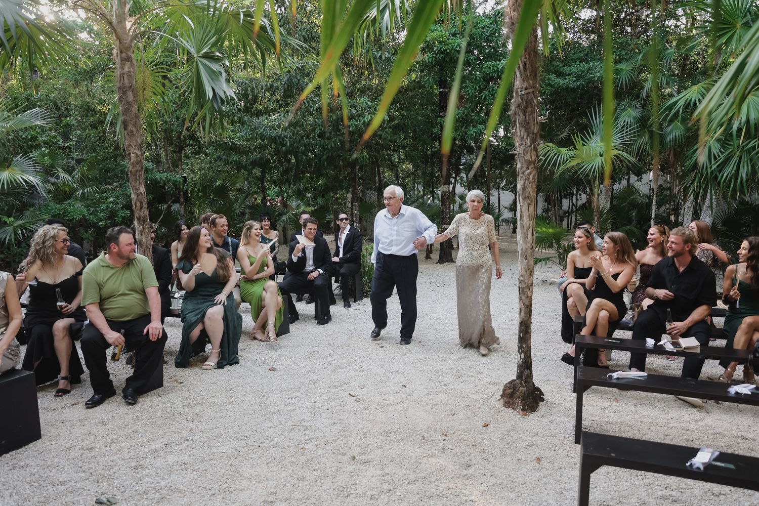 Outdoor wedding ceremony in a tropical garden with guests seated on benches as couple walks down sandy aisle.