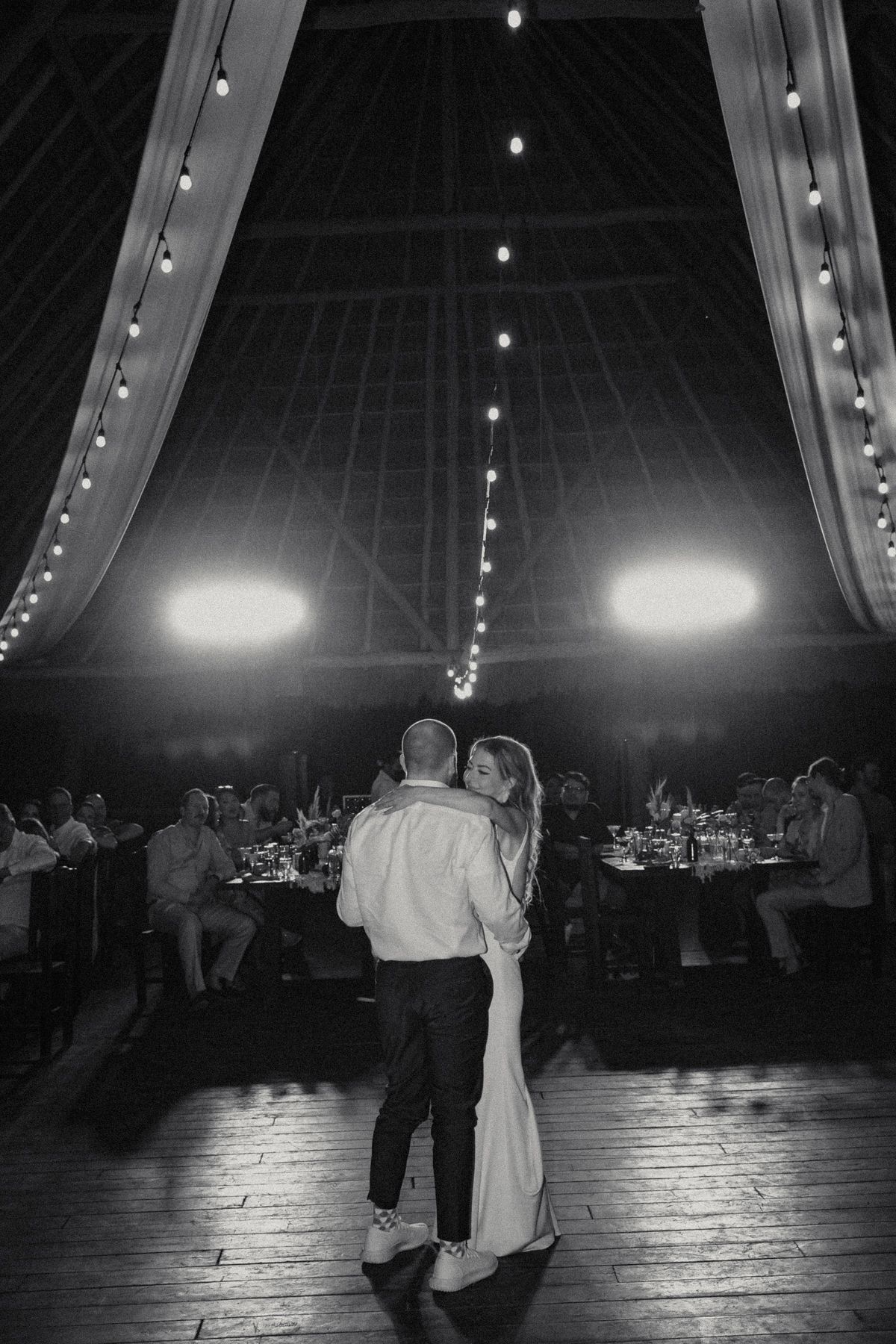 Black and white romantic first dance photo of bride and groom under string lights in venue.