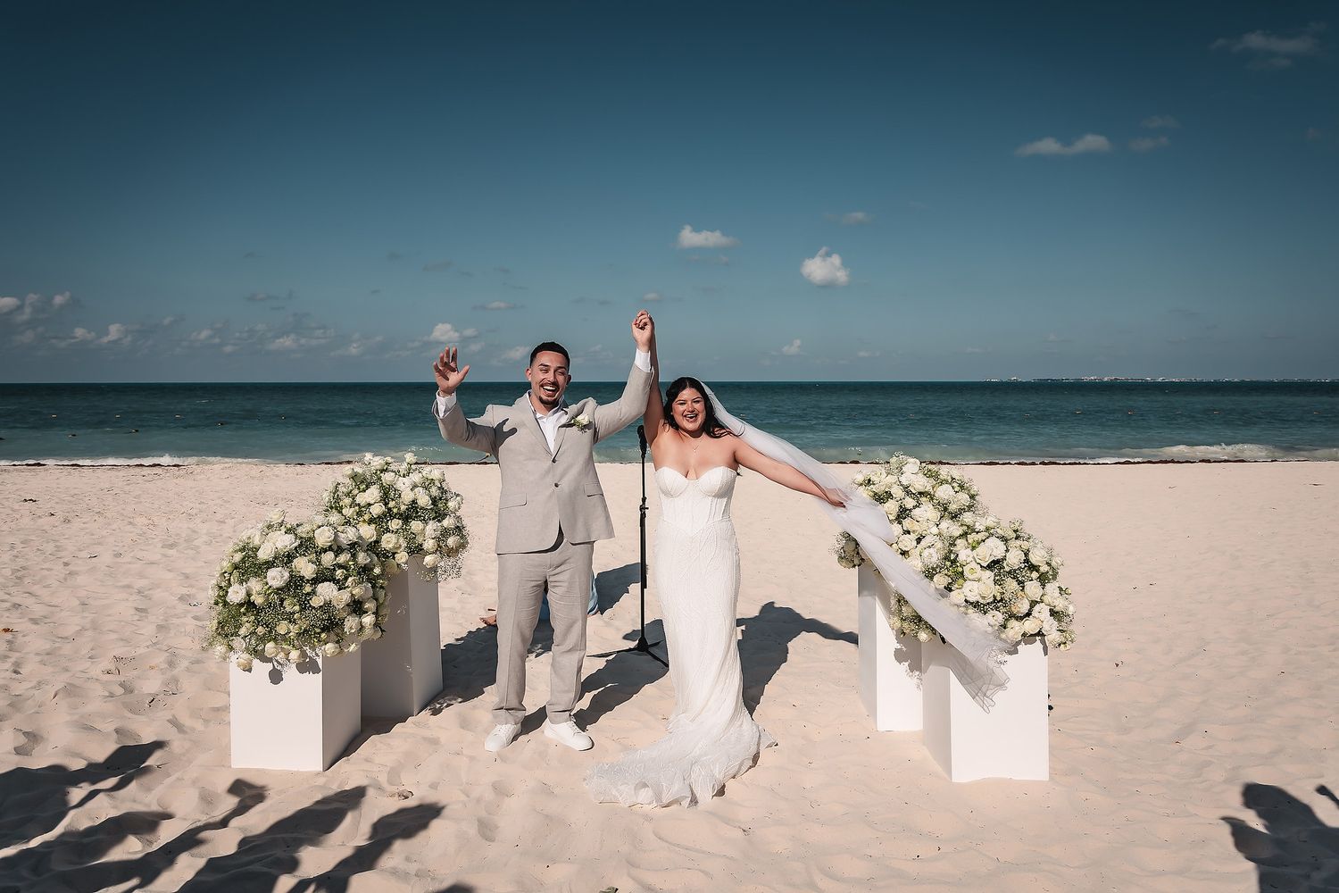 Joyful newlywed couple celebrating on a white sand beach wedding ceremony with white floral arrangements under blue sky.
