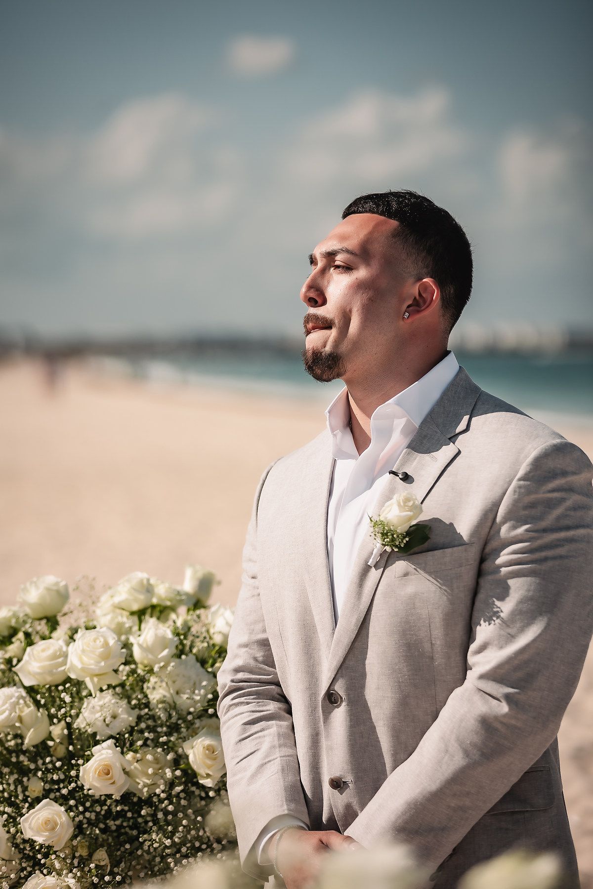 Groom in gray suit with white boutonniere stands at beach wedding altar with white roses.