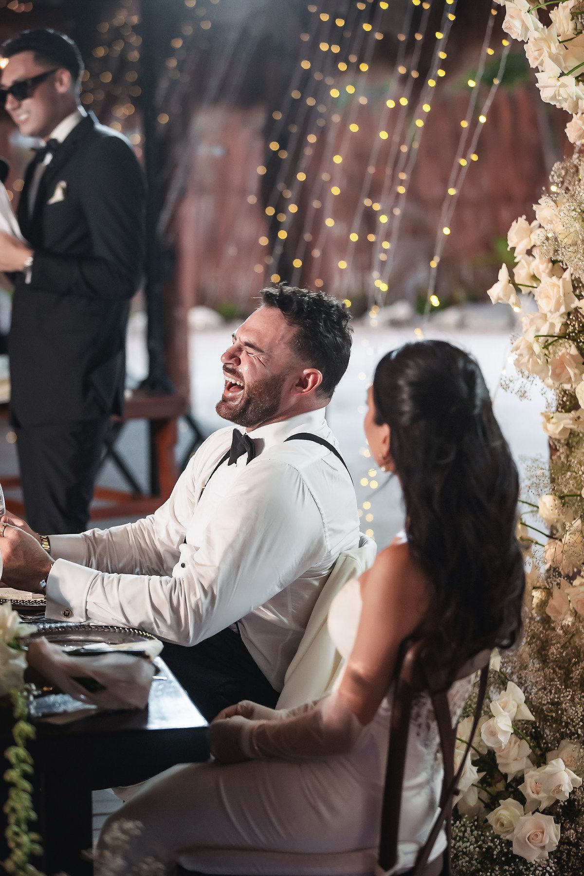 Groom laughing joyfully at wedding reception table beside bride, surrounded by white flowers and fairy lights.