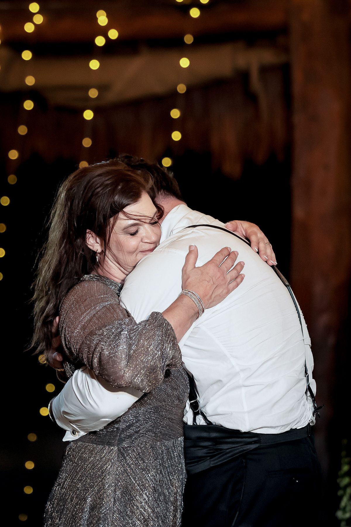 Mother and groom share an emotional hug during a wedding reception dance, with warm bokeh string lights in background.