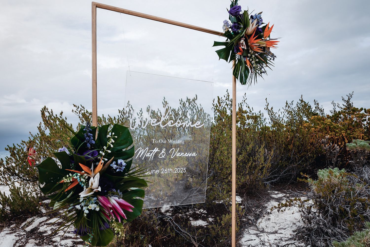 Elegant acrylic wedding welcome sign with gold frame and tropical floral arrangement on a sandy outdoor backdrop.
