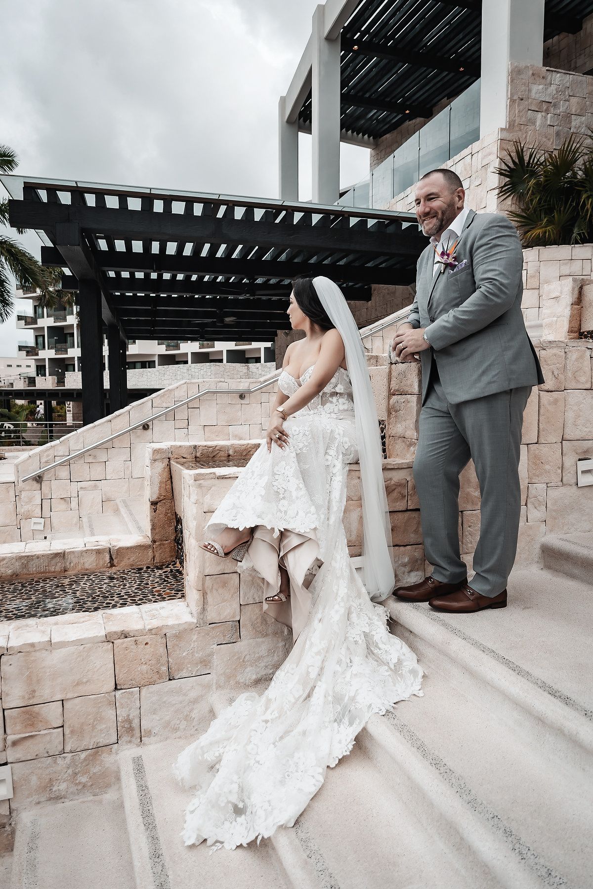 Bride in lace gown and groom in gray suit pose on stone steps at an elegant outdoor wedding venue.