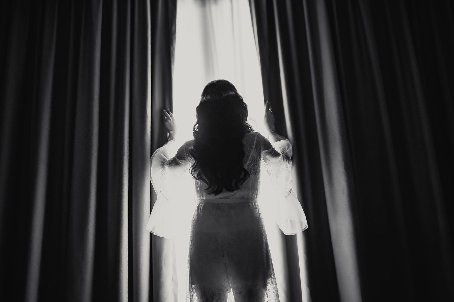Black and white photo of a woman in a lace dress opening curtains, viewed from behind.