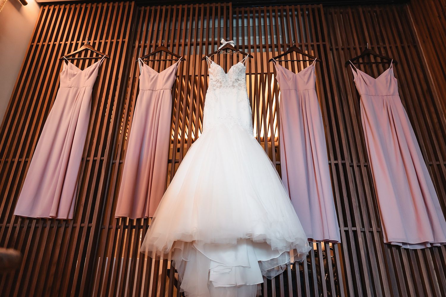 White wedding gown and pink bridesmaid dresses hanging on a wooden slatted wall before the ceremony.