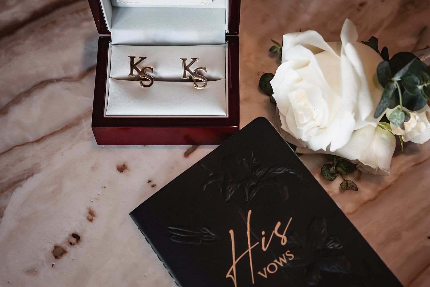 Gold monogram KS cufflinks in a wooden box beside a white rose and a black His Vows wedding book.