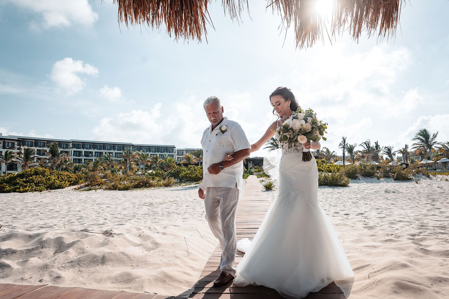 Bride in white gown walks with groom on sandy beach at tropical resort under sunny sky with bouquet.