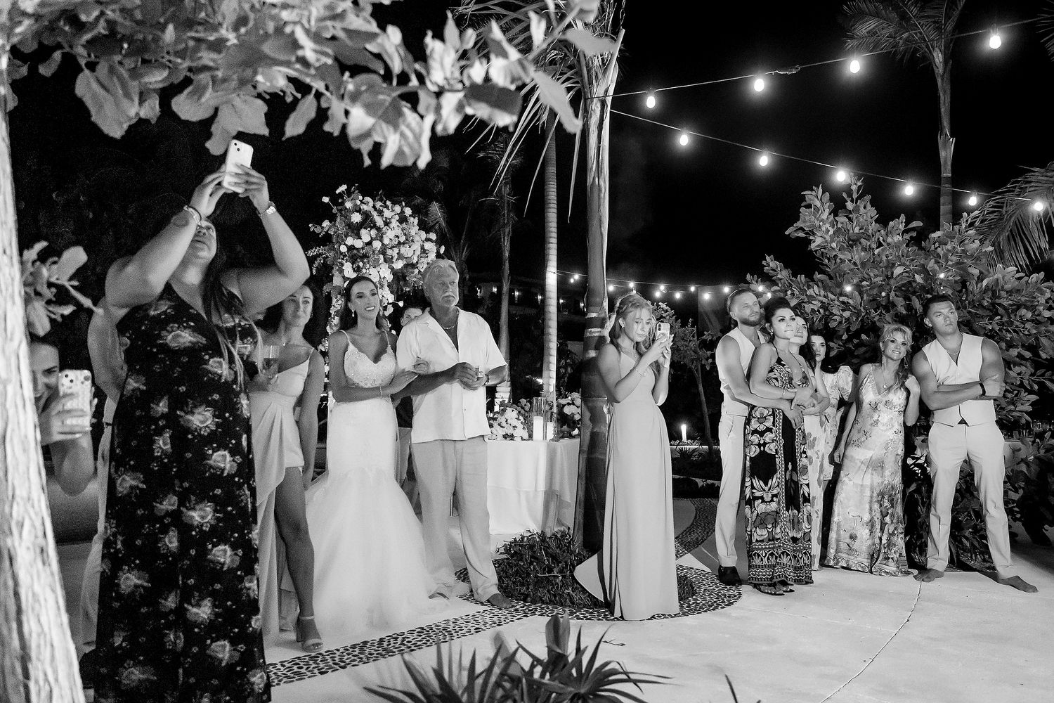 Black and white wedding reception photo with guests celebrating outdoors at night under string lights.