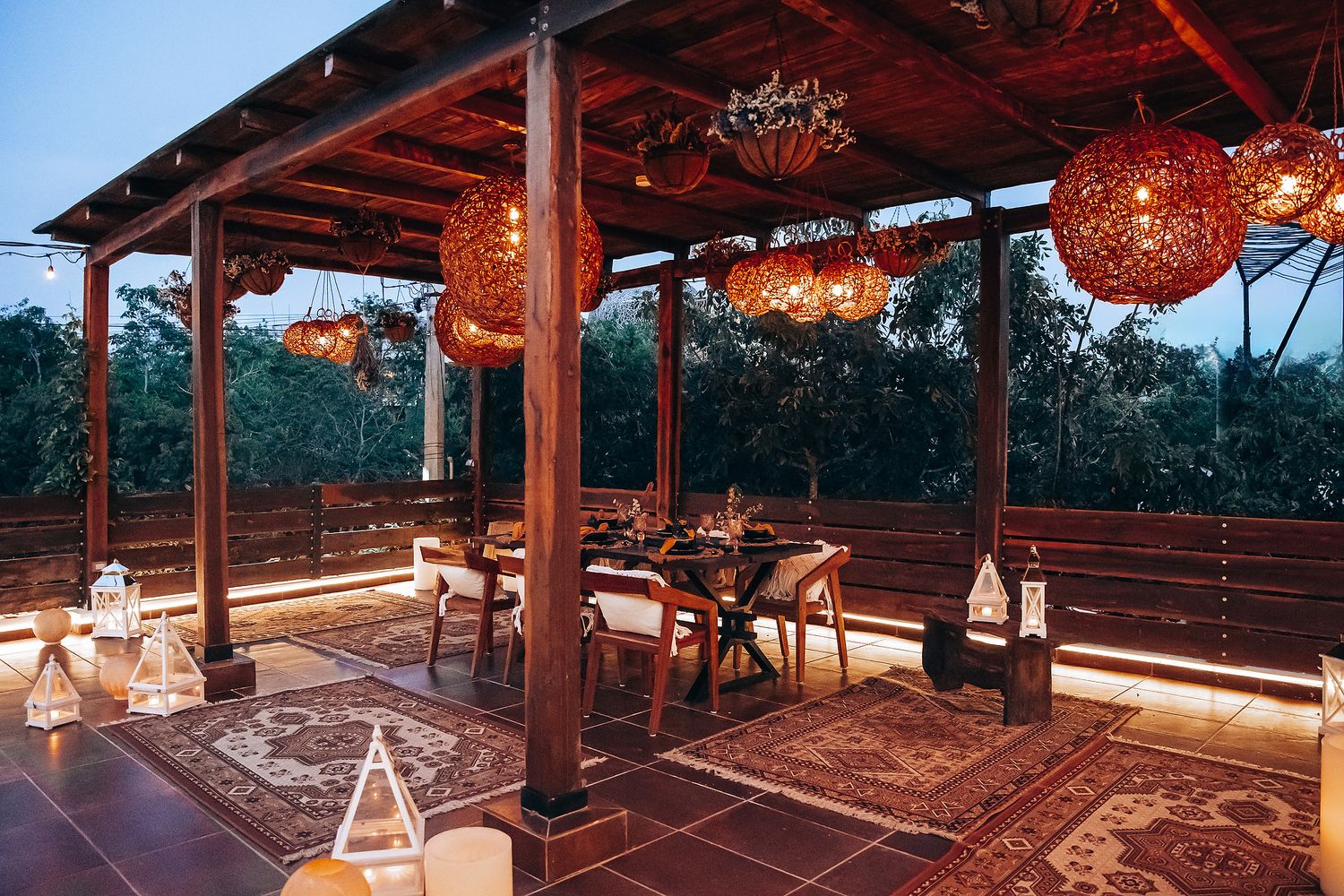 Rustic outdoor wedding dining pavilion with woven pendant lights and patterned rugs at dusk.