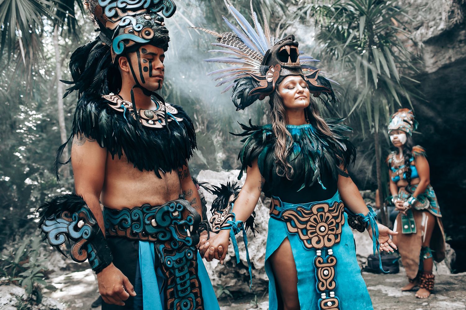 Two performers in vibrant Mayan ceremonial attire with feathered headdresses walk hand in hand through a jungle cave.