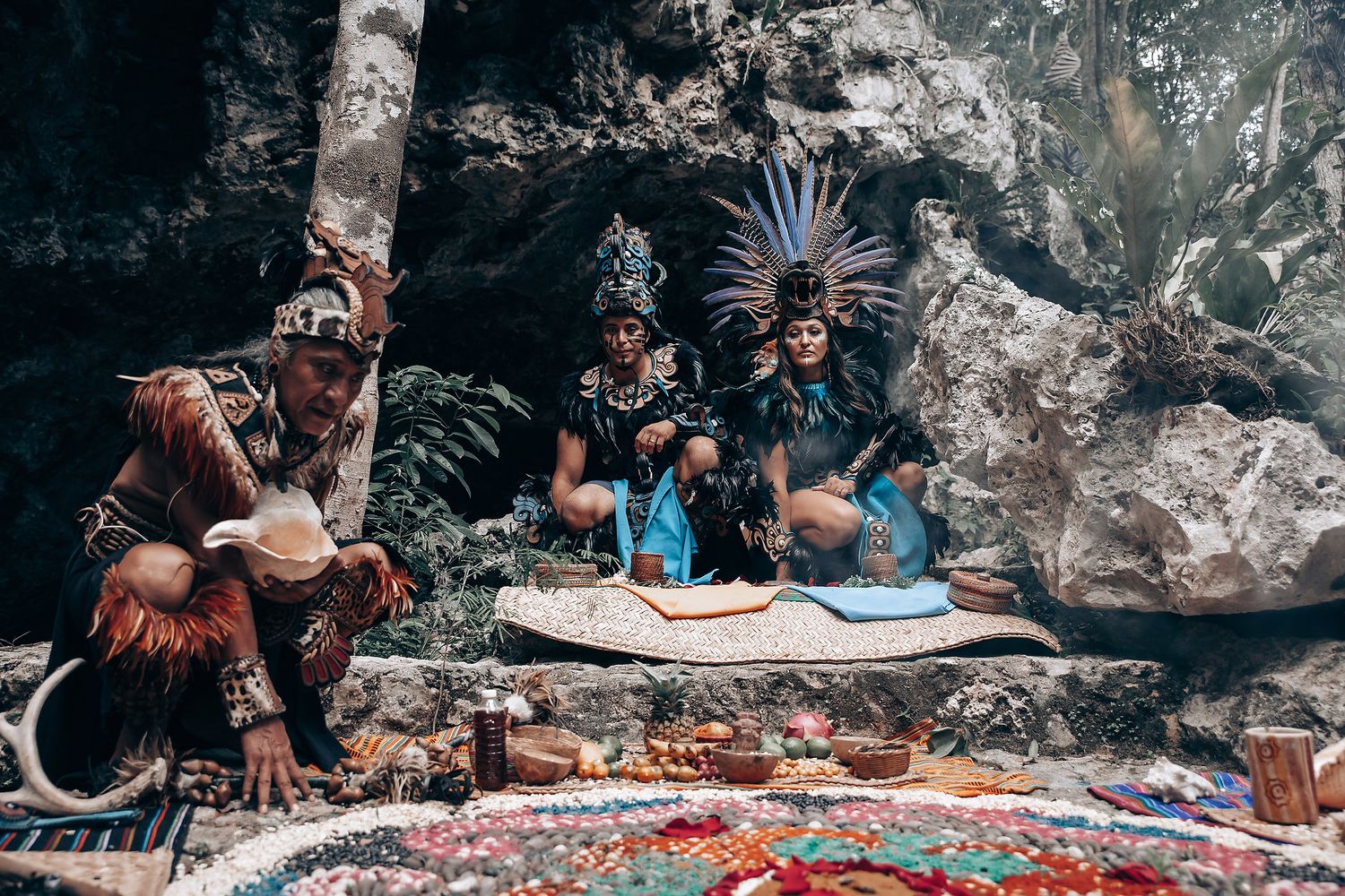 Three people in elaborate Mayan ceremonial dress sit surrounded by colorful offerings and artifacts in a cave setting.