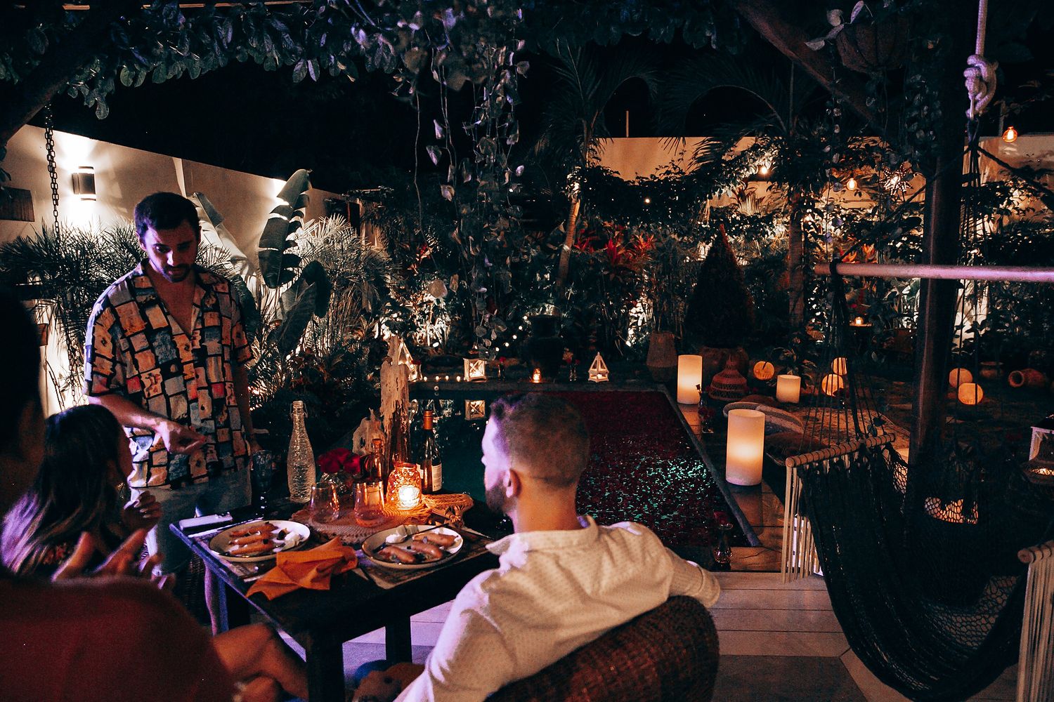 Guests enjoy an intimate candlelit outdoor dinner surrounded by lush greenery and warm ambient lighting at night.