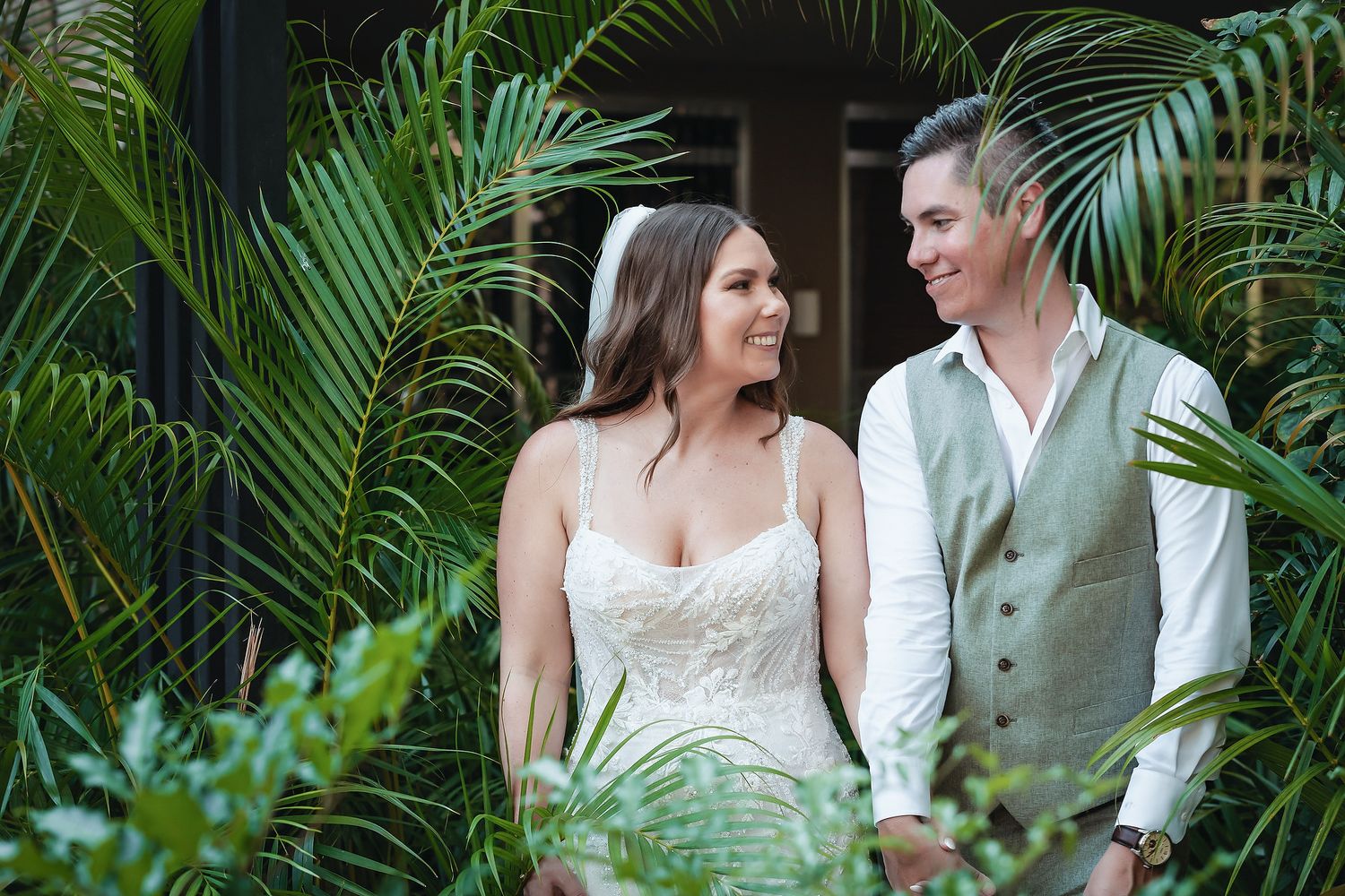 Bride and groom smiling at each other surrounded by lush tropical palm leaves outdoors.