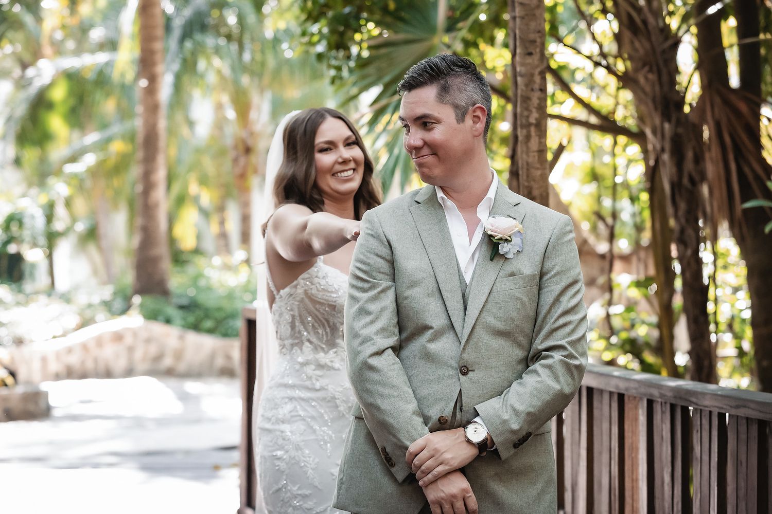 Bride in white lace gown taps groom in gray suit on shoulder during outdoor wedding first look among tropical trees.