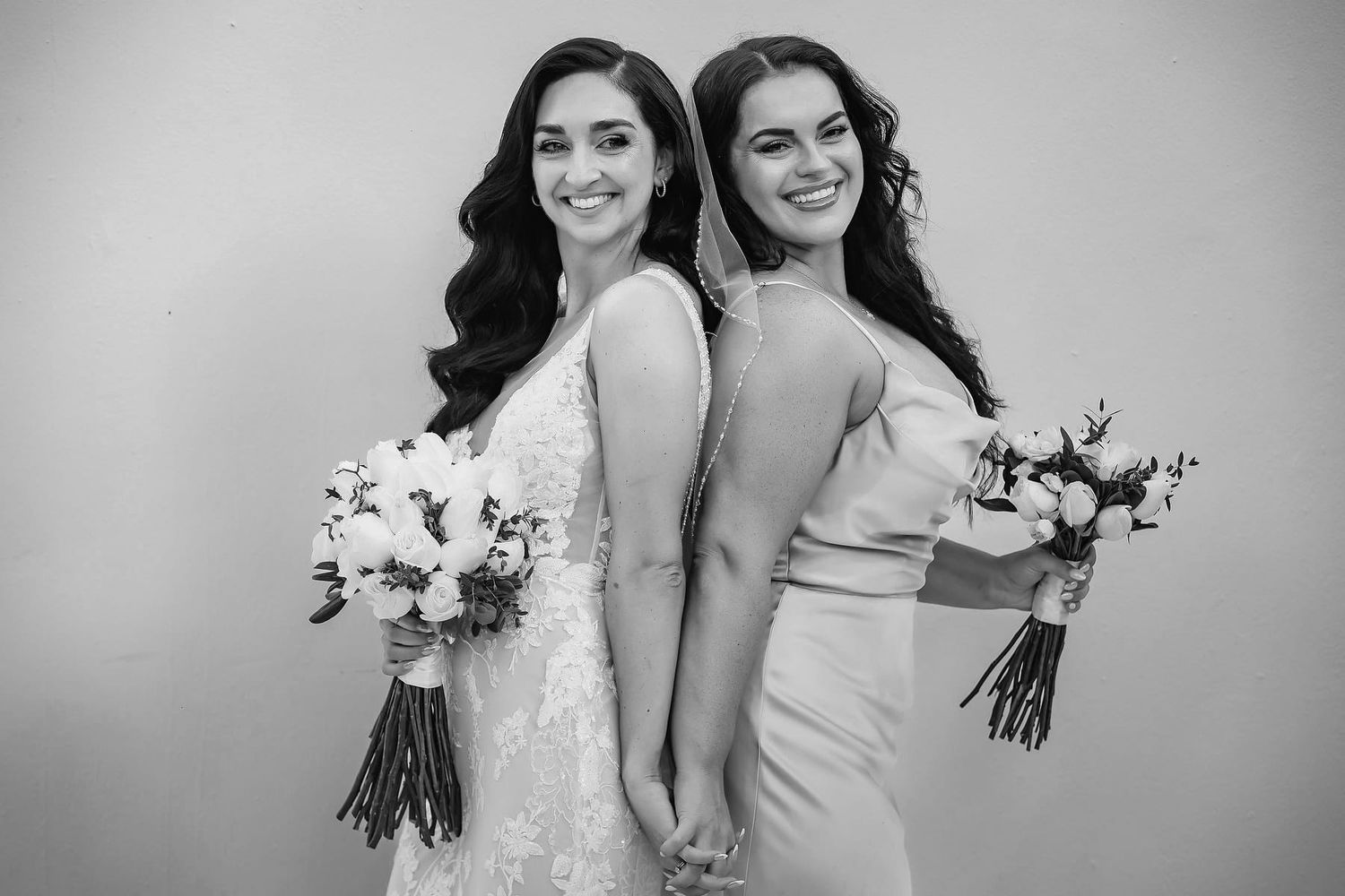 Two people in white dresses stand close together while holding bouquets in black and white photography.
