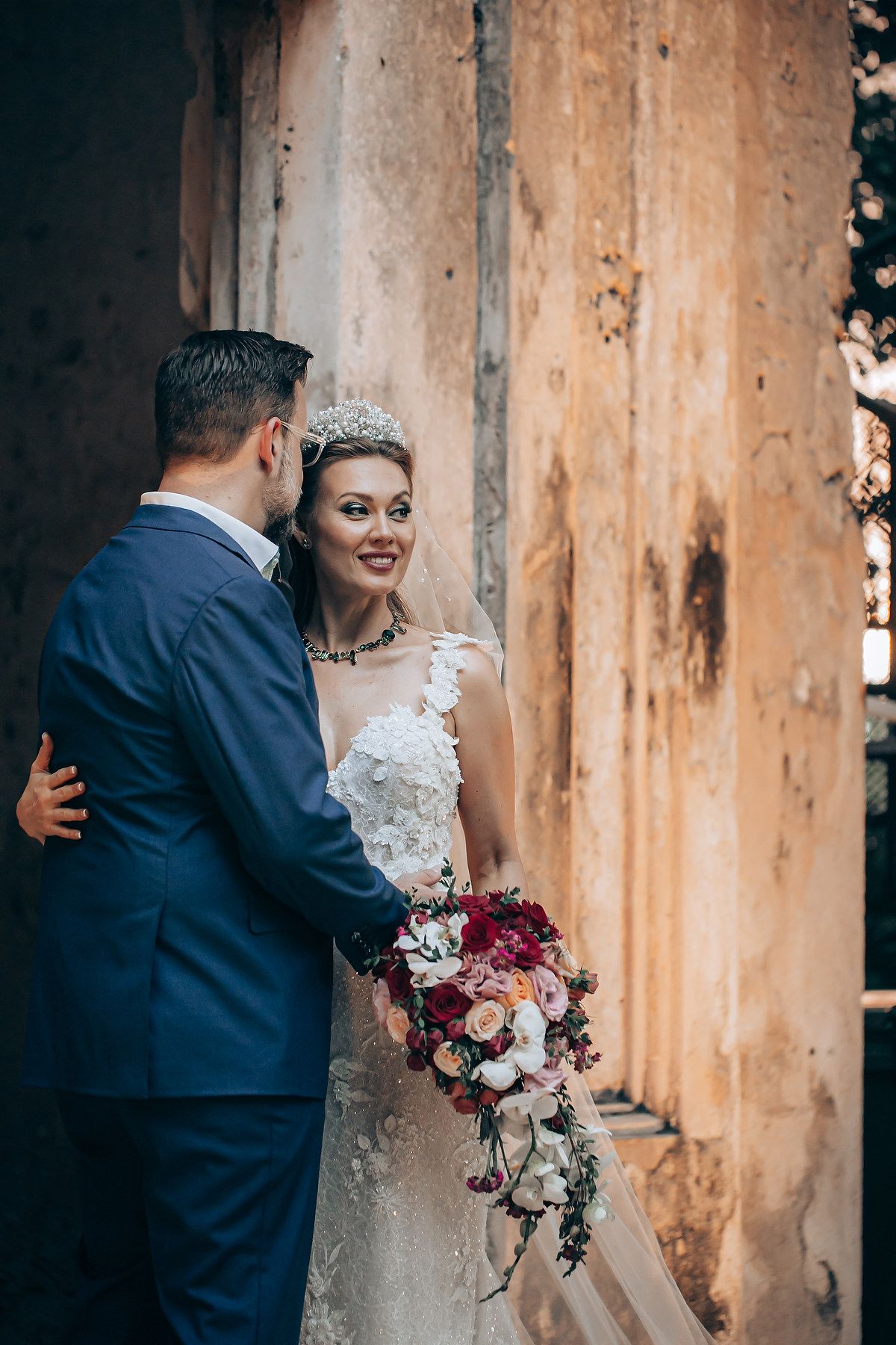 A romantic wedding couple embraces against a rustic wooden wall while holding a burgundy and white floral bouquet.