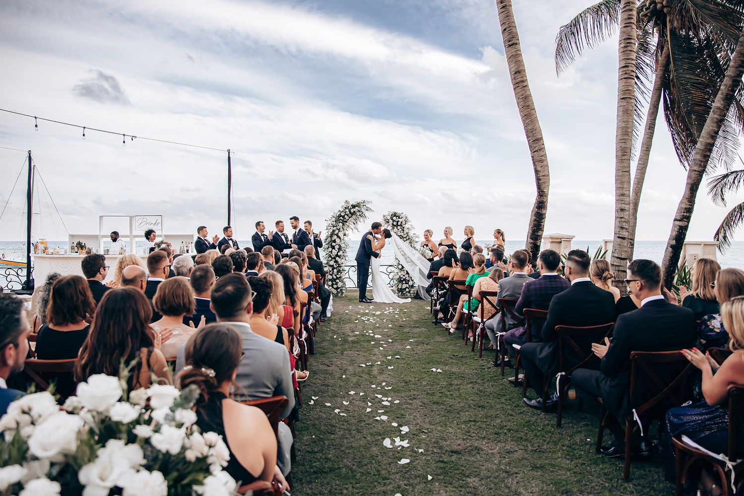 Outdoor beach wedding ceremony with palm trees and white floral decorations along aisle filled with seated guests.