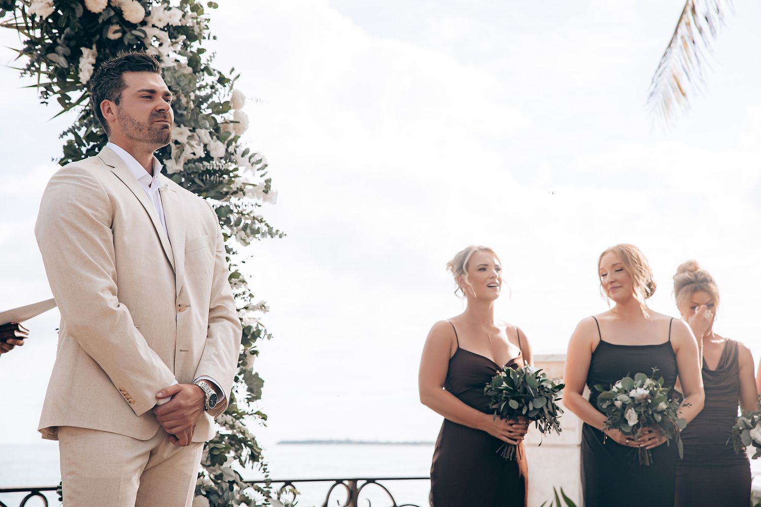 Wedding party stands at outdoor ceremony with groom in beige suit and bridesmaids in black dresses.