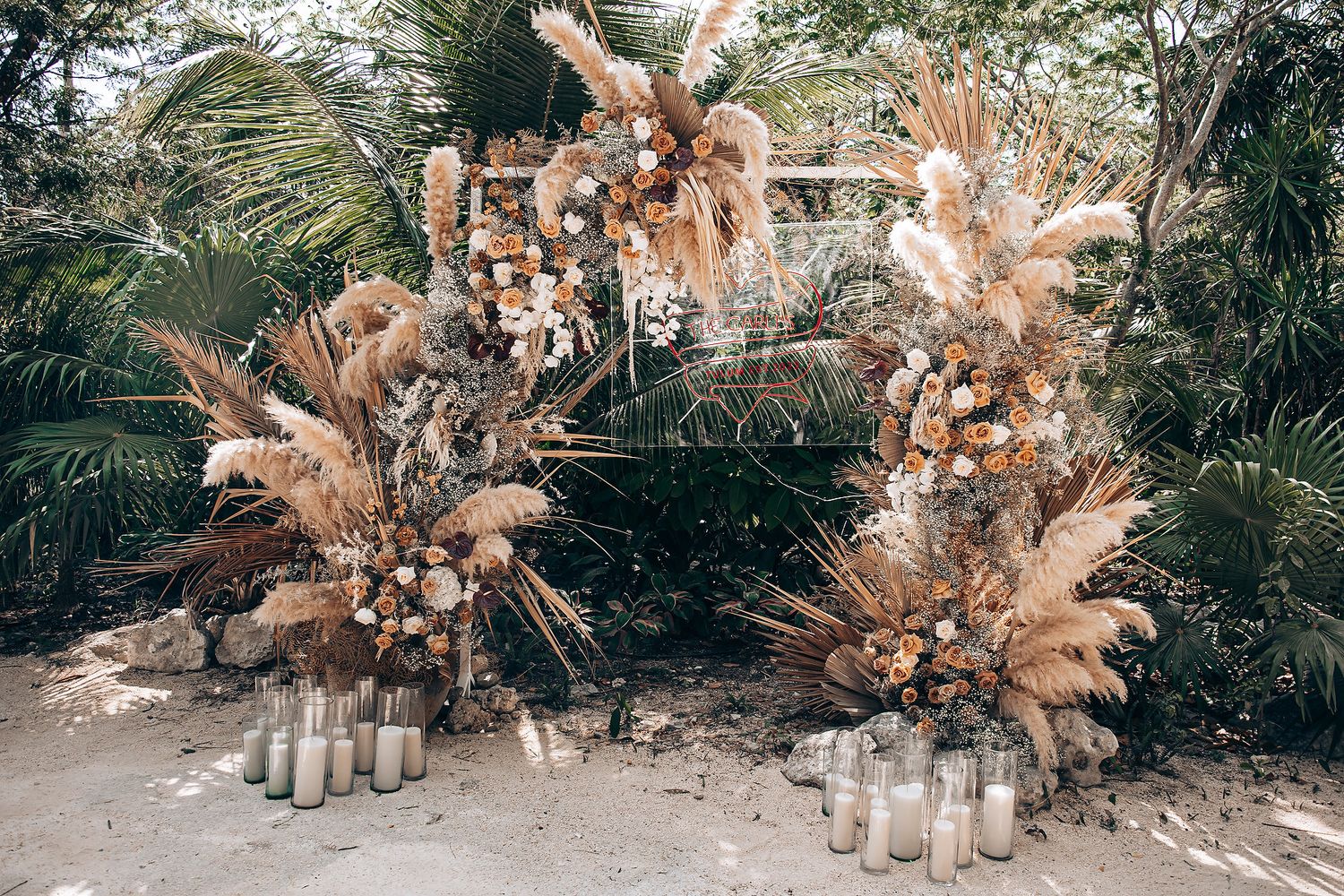 Natural boho wedding decor featuring dried palm fronds, pampas grass, and candles arranged along a sandy pathway.