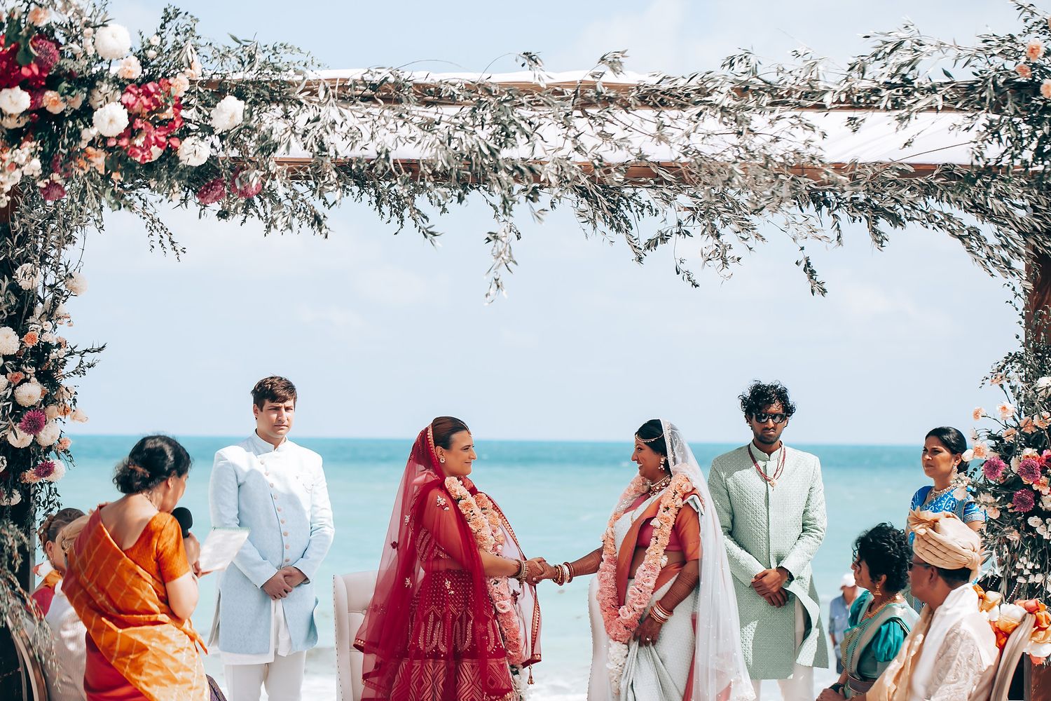 Wedding guests in vibrant saris and formal wear mingle at an outdoor beach celebration.