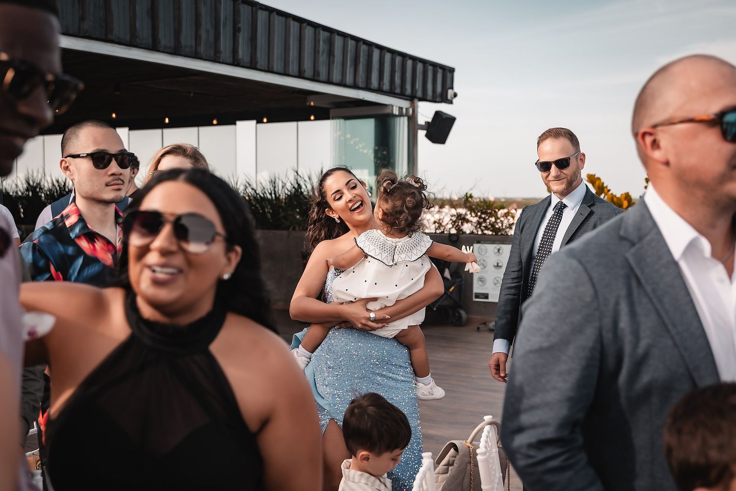 Wedding attendees gather on a sunny outdoor terrace overlooking the ocean.