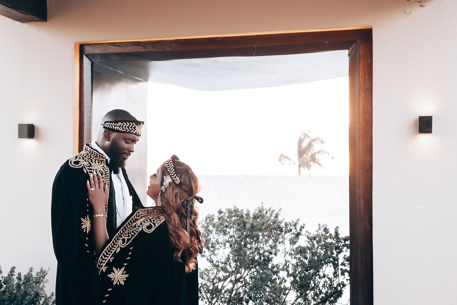 A couple in wedding attire embraces while standing in front of a scenic outdoor view at sunset.