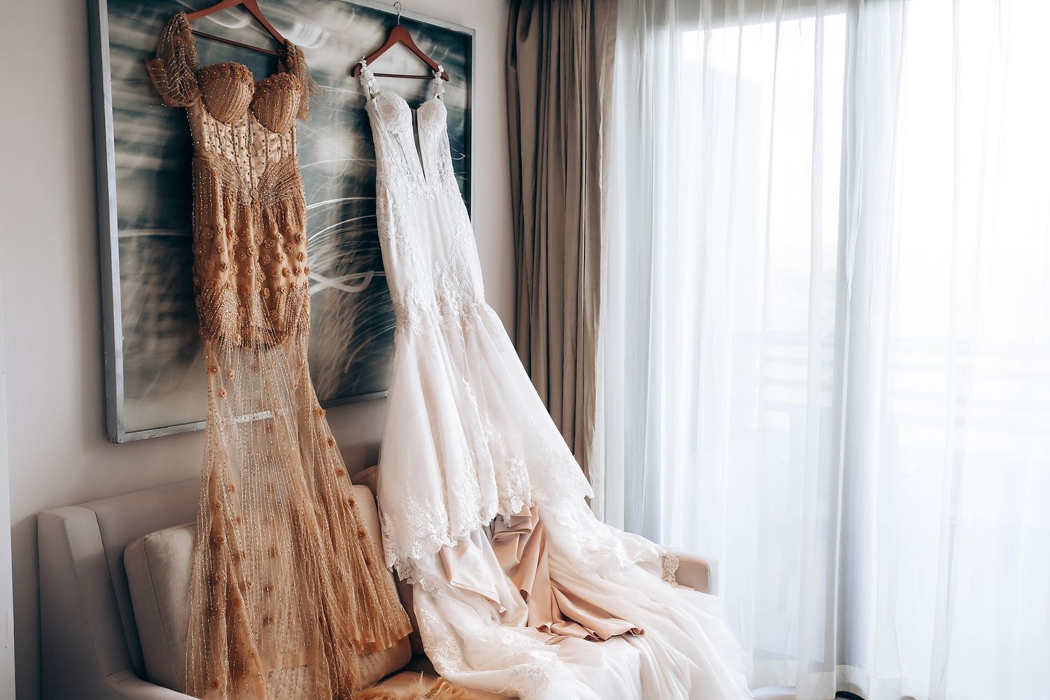 Elegant wedding dresses hang on display against a white curtained wall with natural lighting.