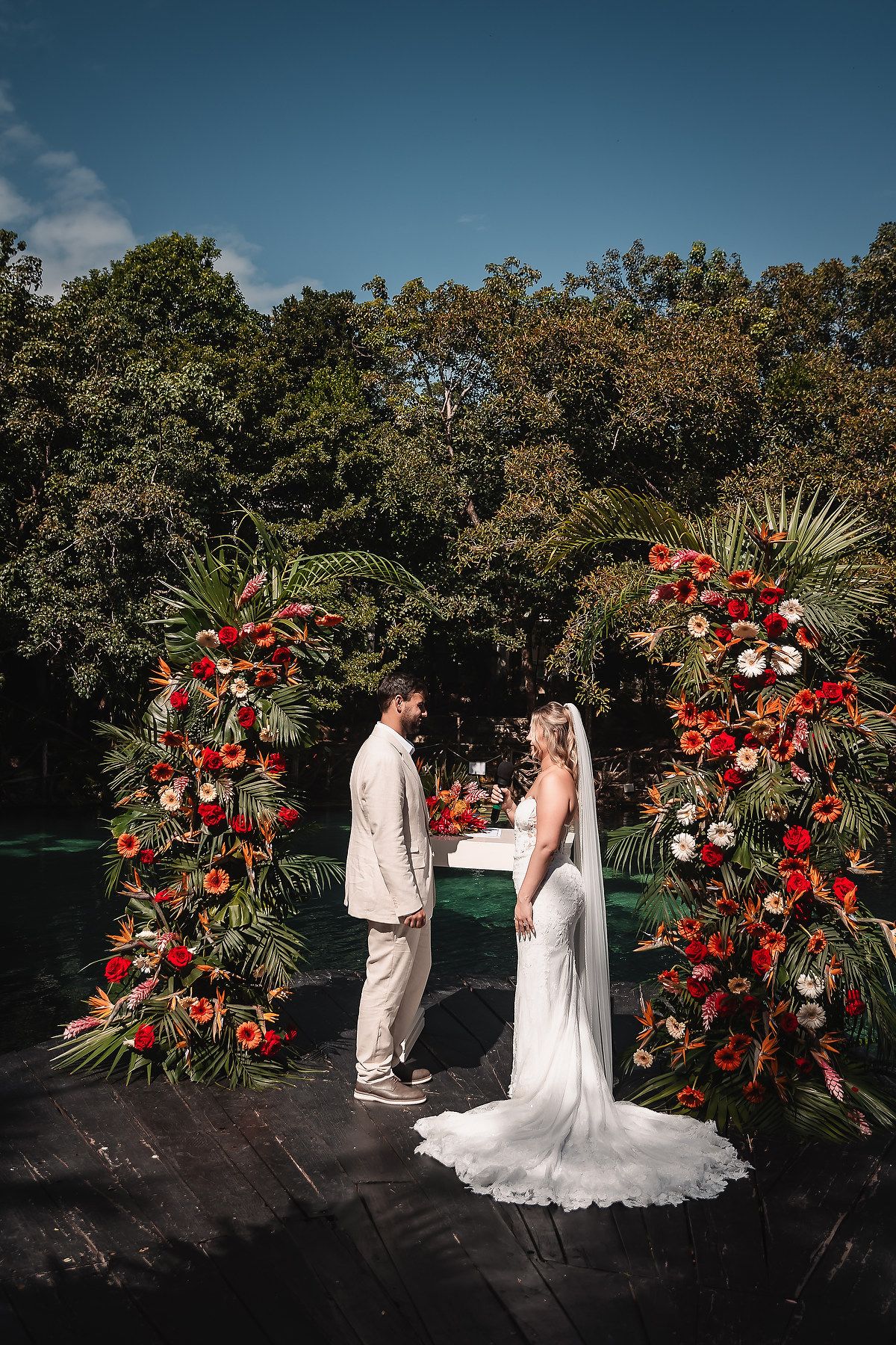 Wedding ceremony at a tropical venue with dramatic red floral arches against a natural backdrop.