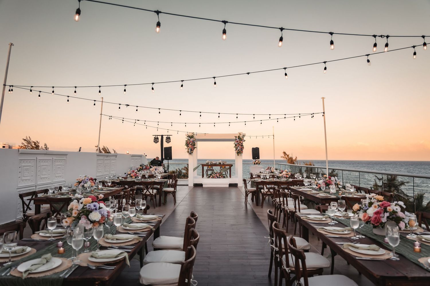 Outdoor reception setup with string lights and ocean views during sunset.