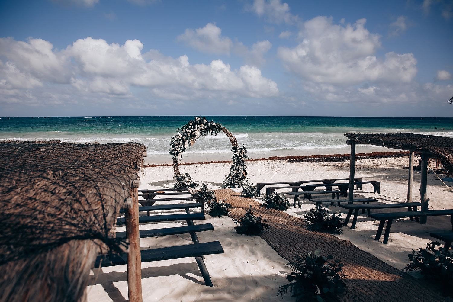 Beachfront wedding ceremony setup with floral arch and wooden benches overlooking turquoise ocean waters.