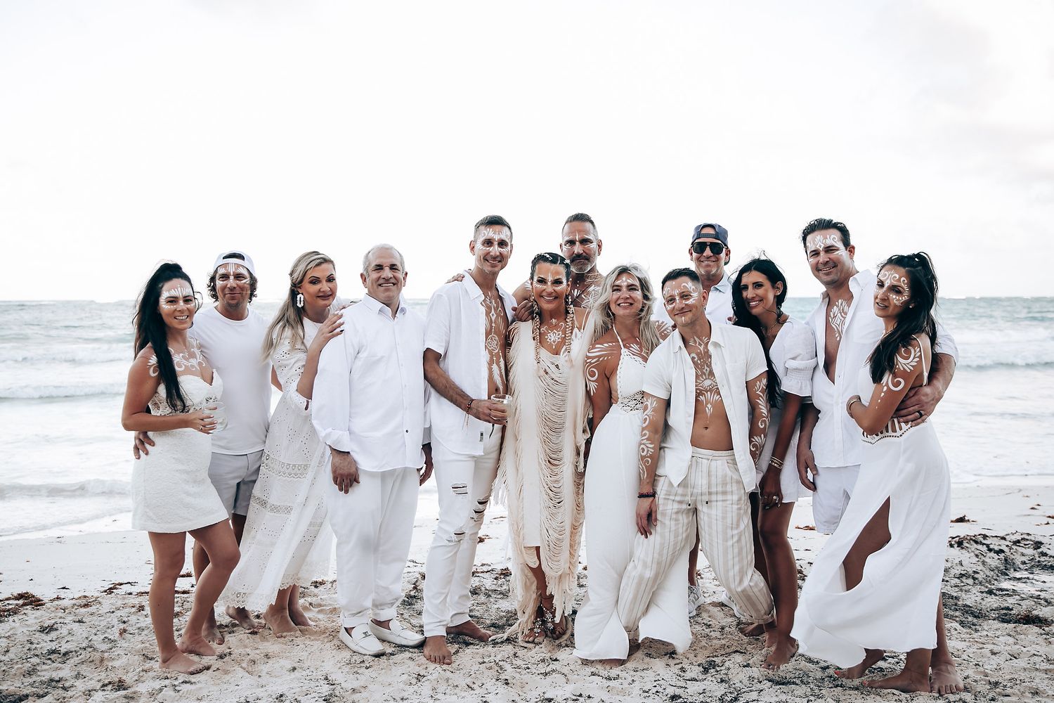 A group photo of wedding guests dressed in white posing together on a beach.