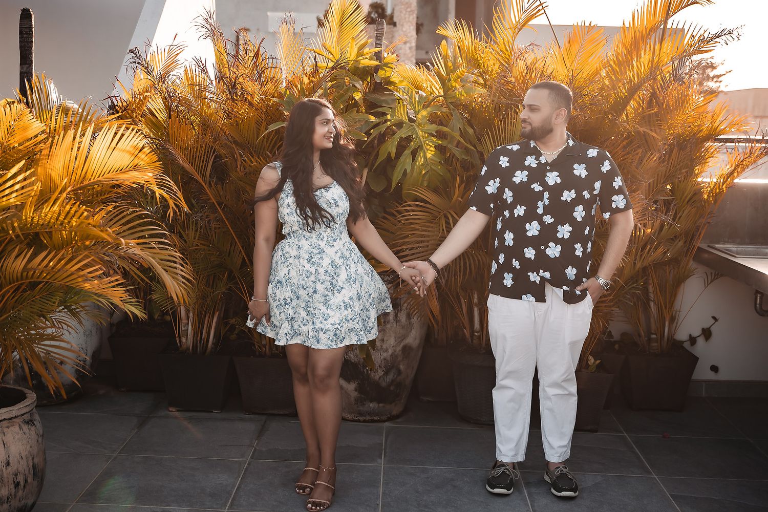 Couple poses in front of lush orange-lit palm garden during golden hour.