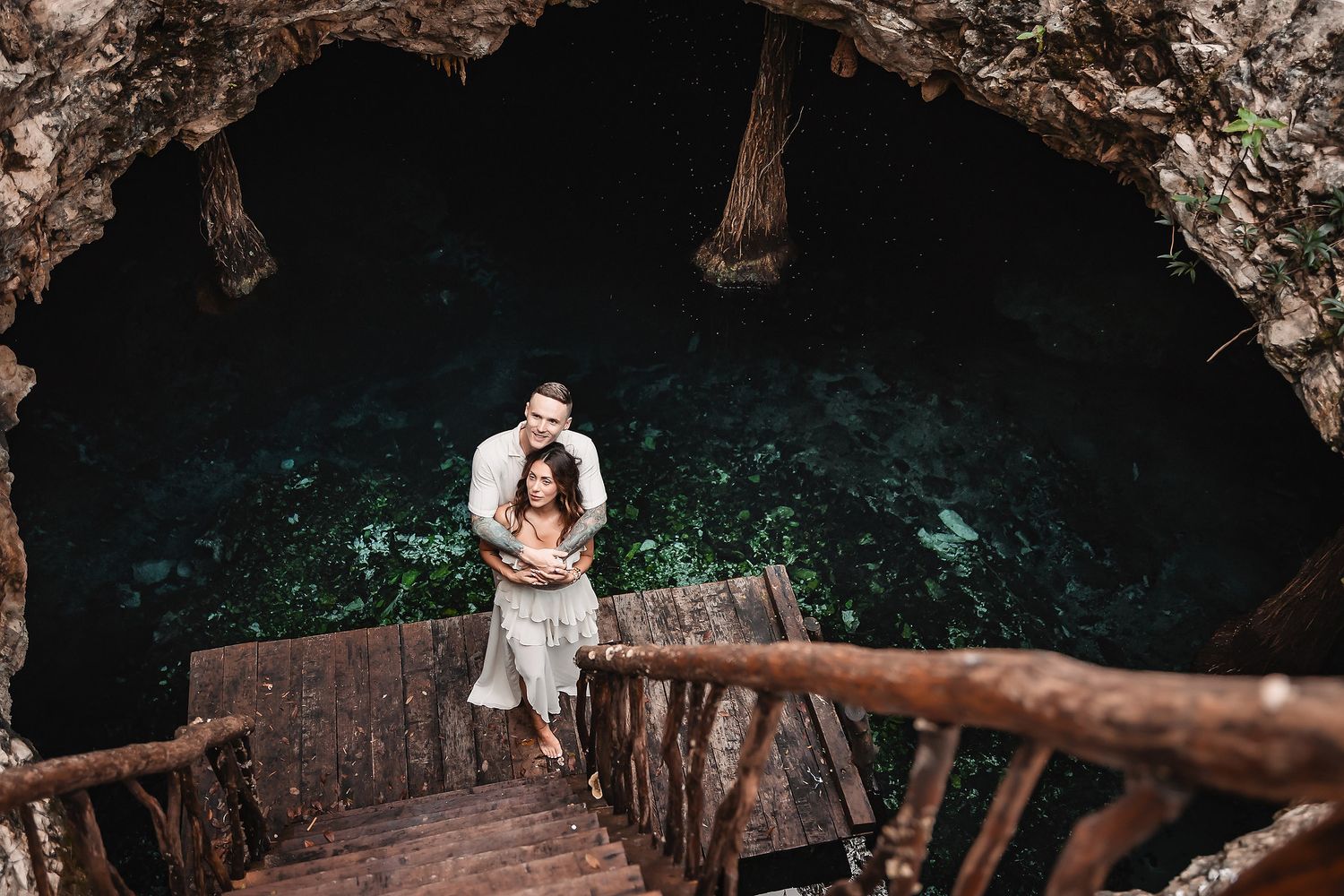 Two people pose on rustic wooden stairs leading down into a mysterious cave.
