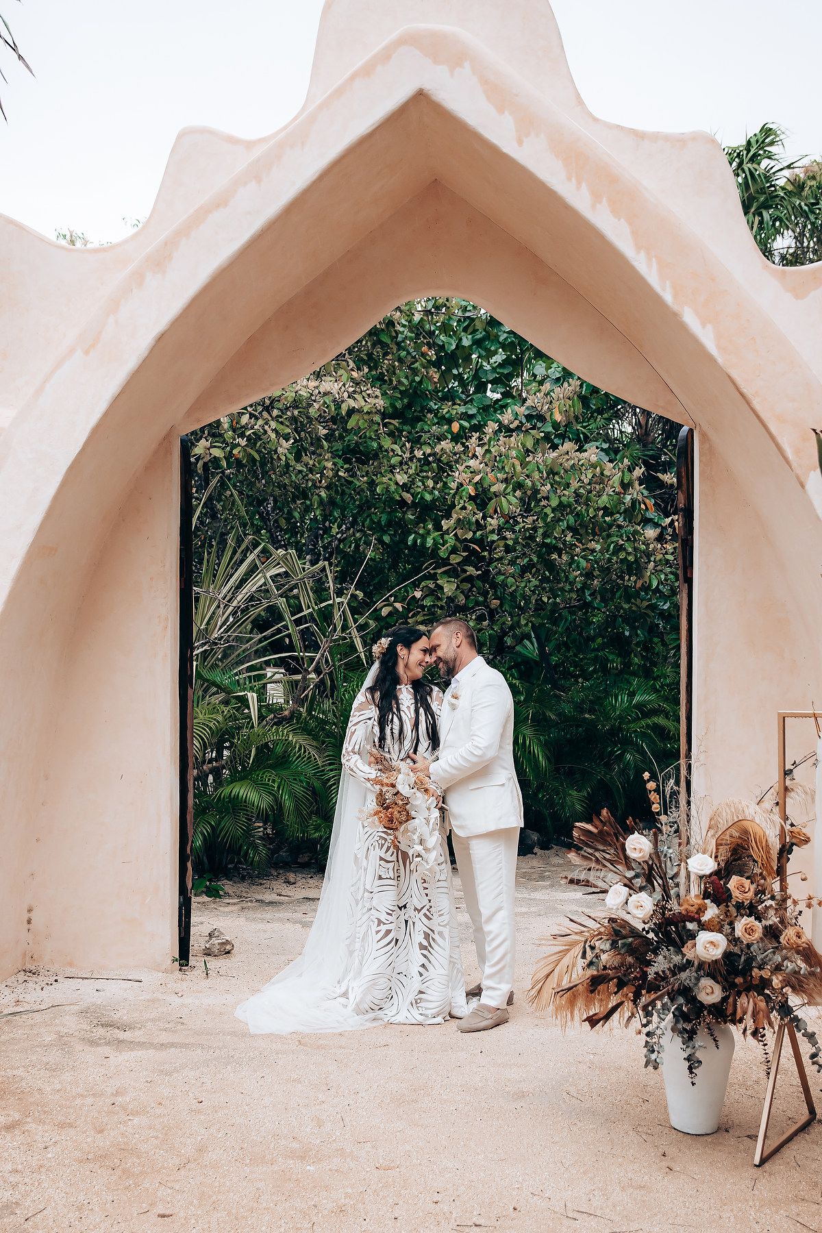 A couple poses under a pink archway with dried floral arrangements and tropical greenery.