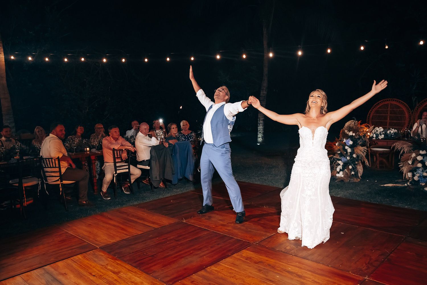 Wedding celebration on outdoor dance floor illuminated by string lights at night.
