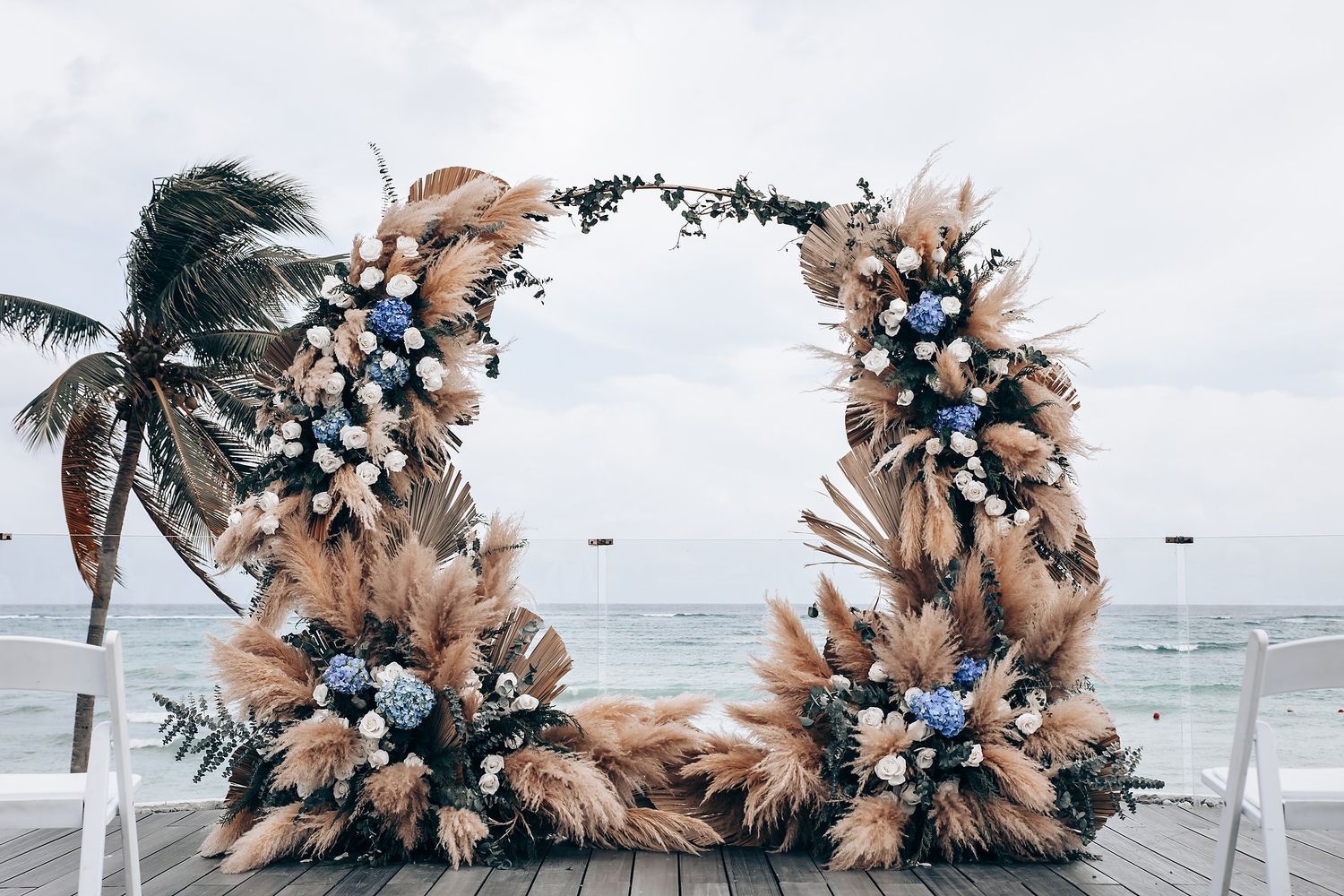 Beachside wedding arch decorated with pampas grass and blue flowers against cloudy sky.