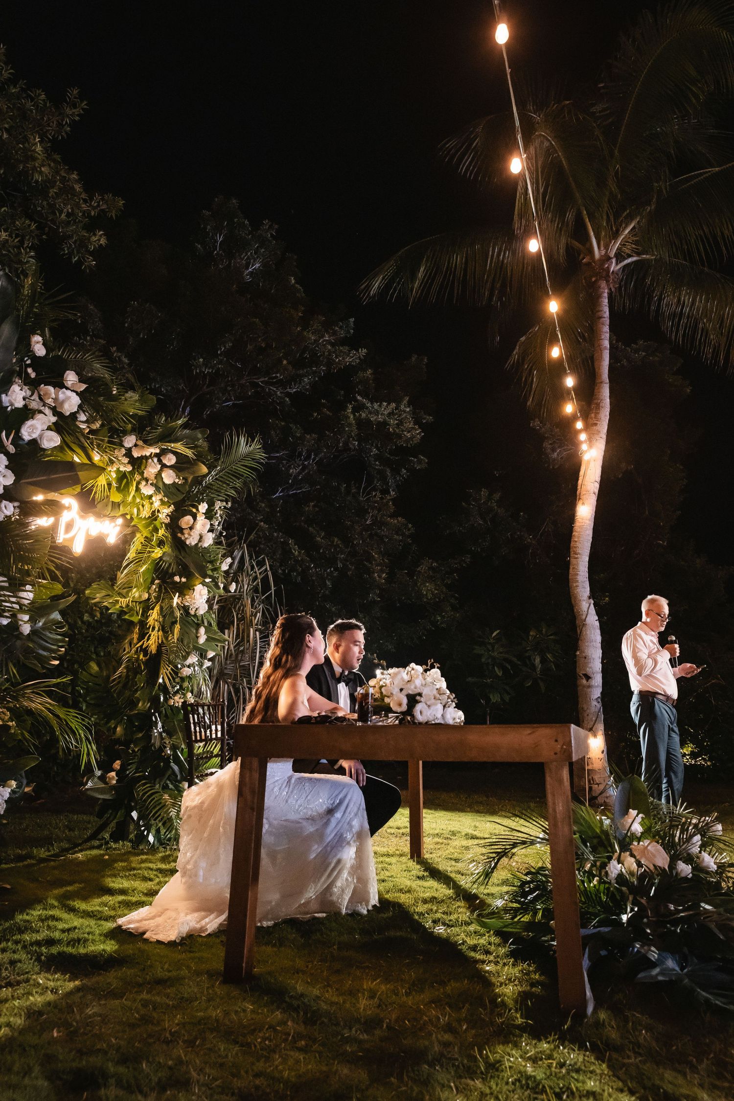 Evening wedding reception seating area with string lights and tropical landscaping.
