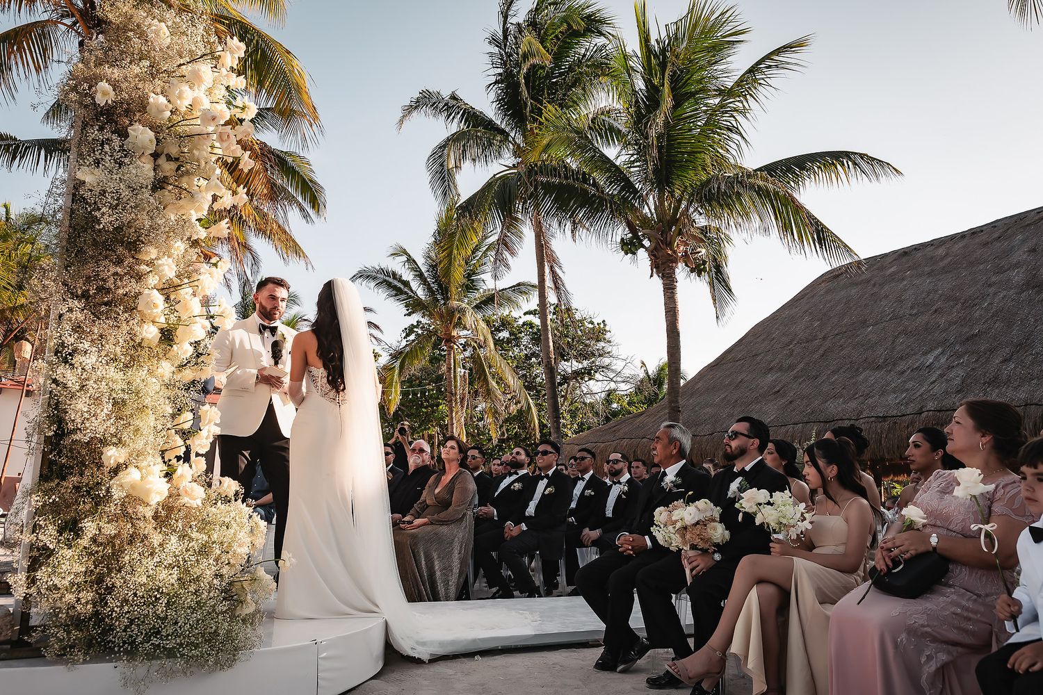 Outdoor wedding ceremony with floral-adorned arch and guests seated on white chairs by palm trees.