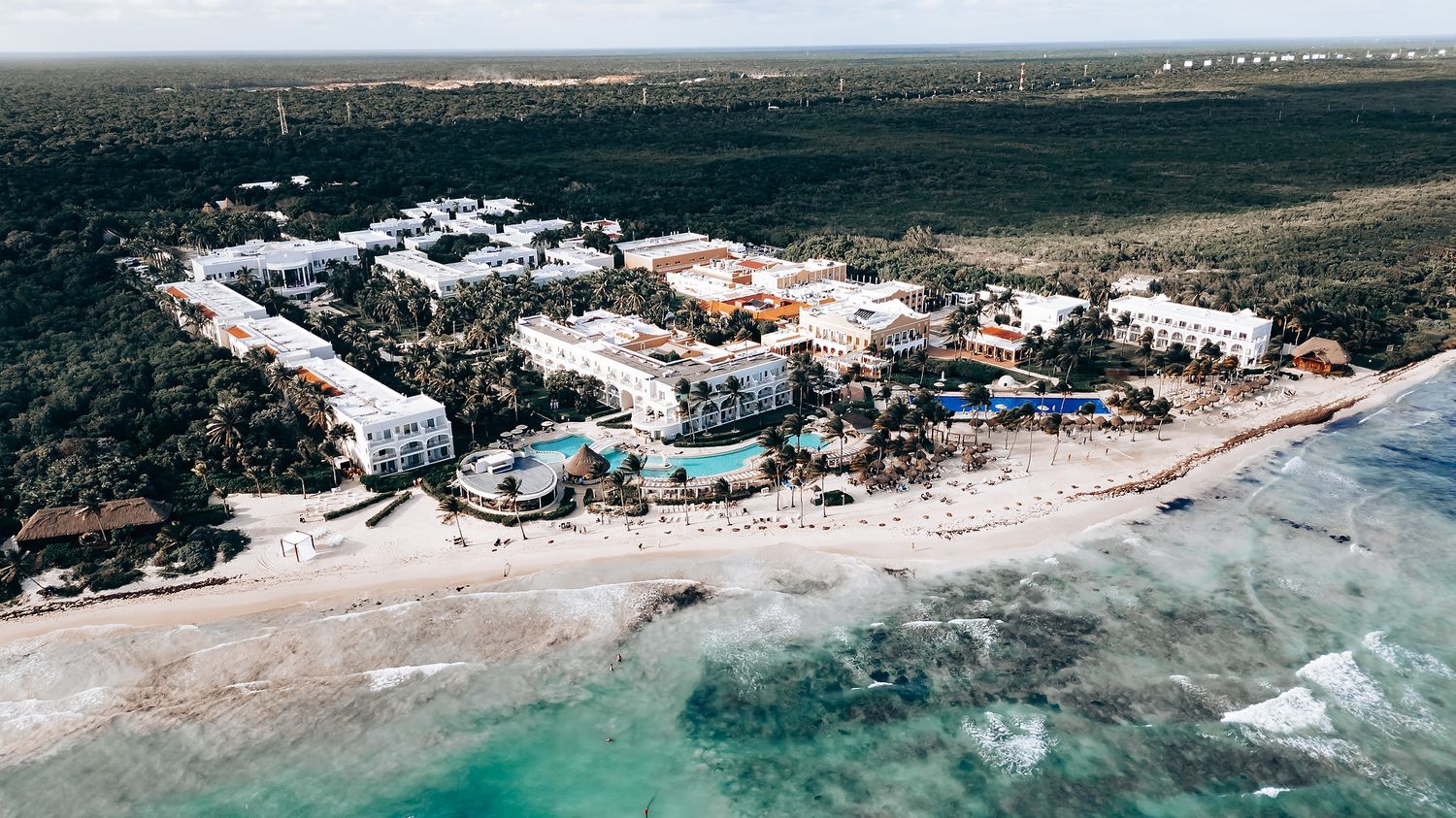 Panoramic drone shot of beachfront properties along tropical coastline with crystal clear waters.