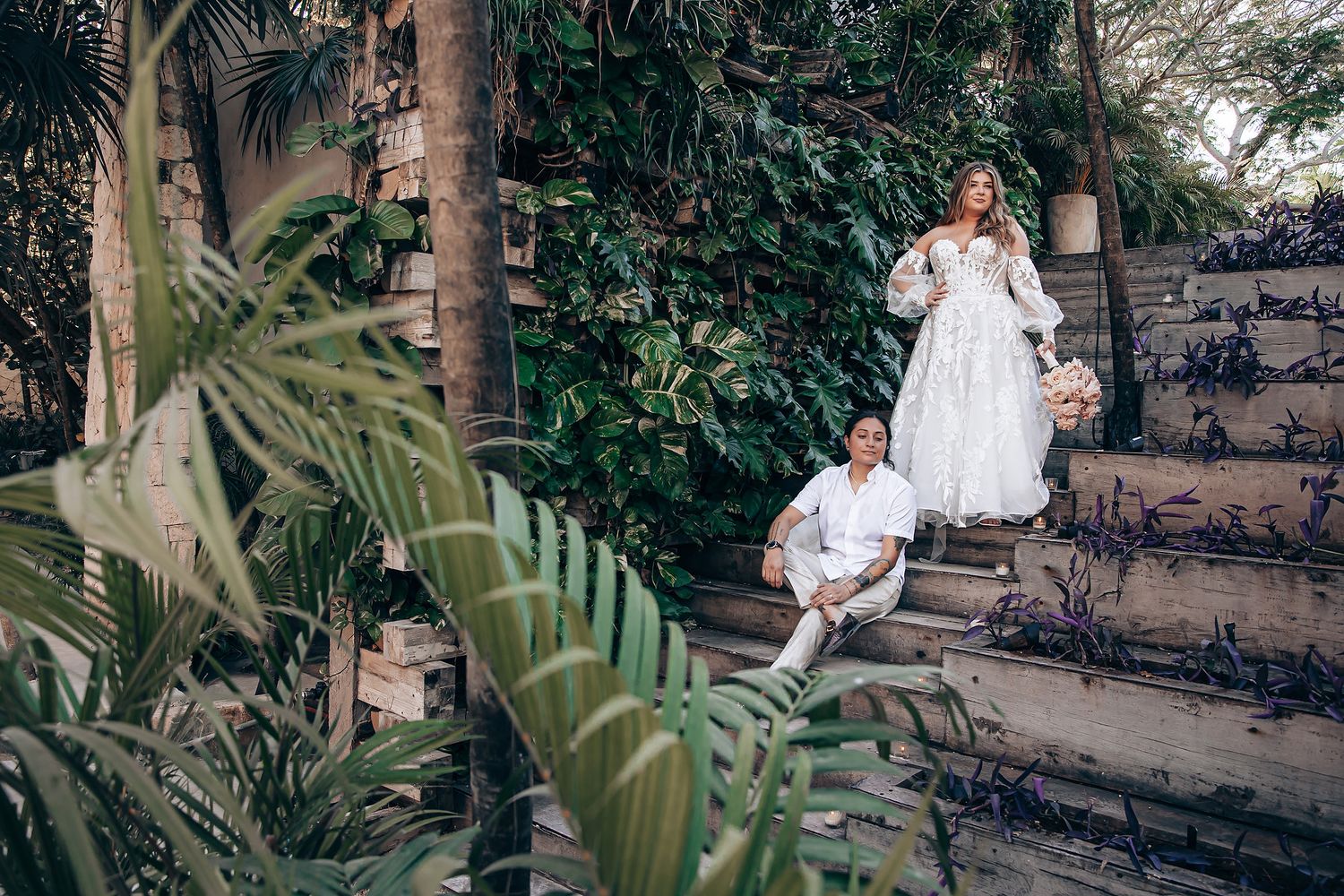 A couple poses in a lush tropical garden setting with stone walls and palm fronds creating a romantic atmosphere.