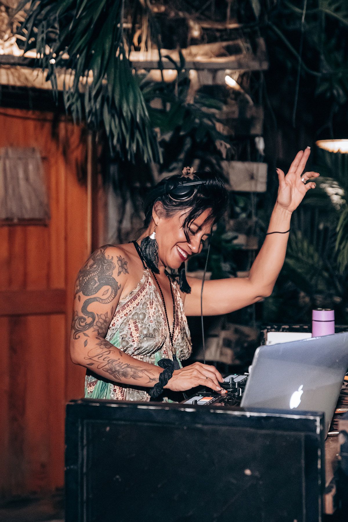 DJ performs at a tropical venue with laptop and mixing equipment while raising hand during energetic set.