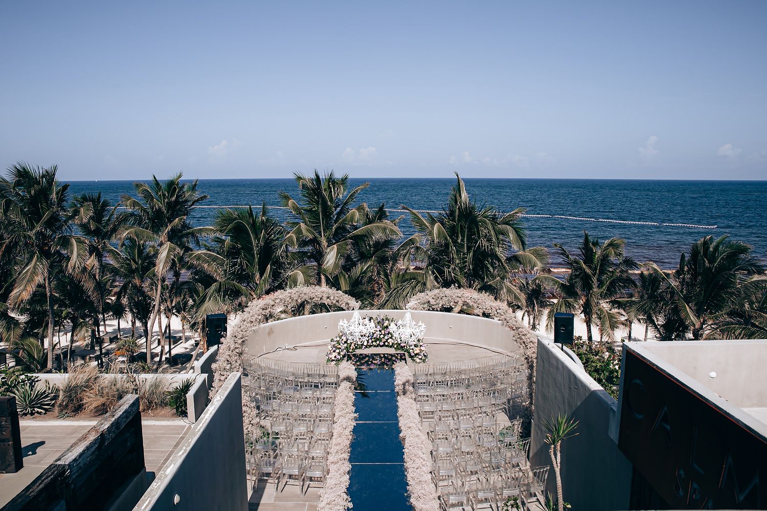 Aerial view of a beachfront resort with palm trees, thatched roofs, and a blue swimming pool leading to the ocean.