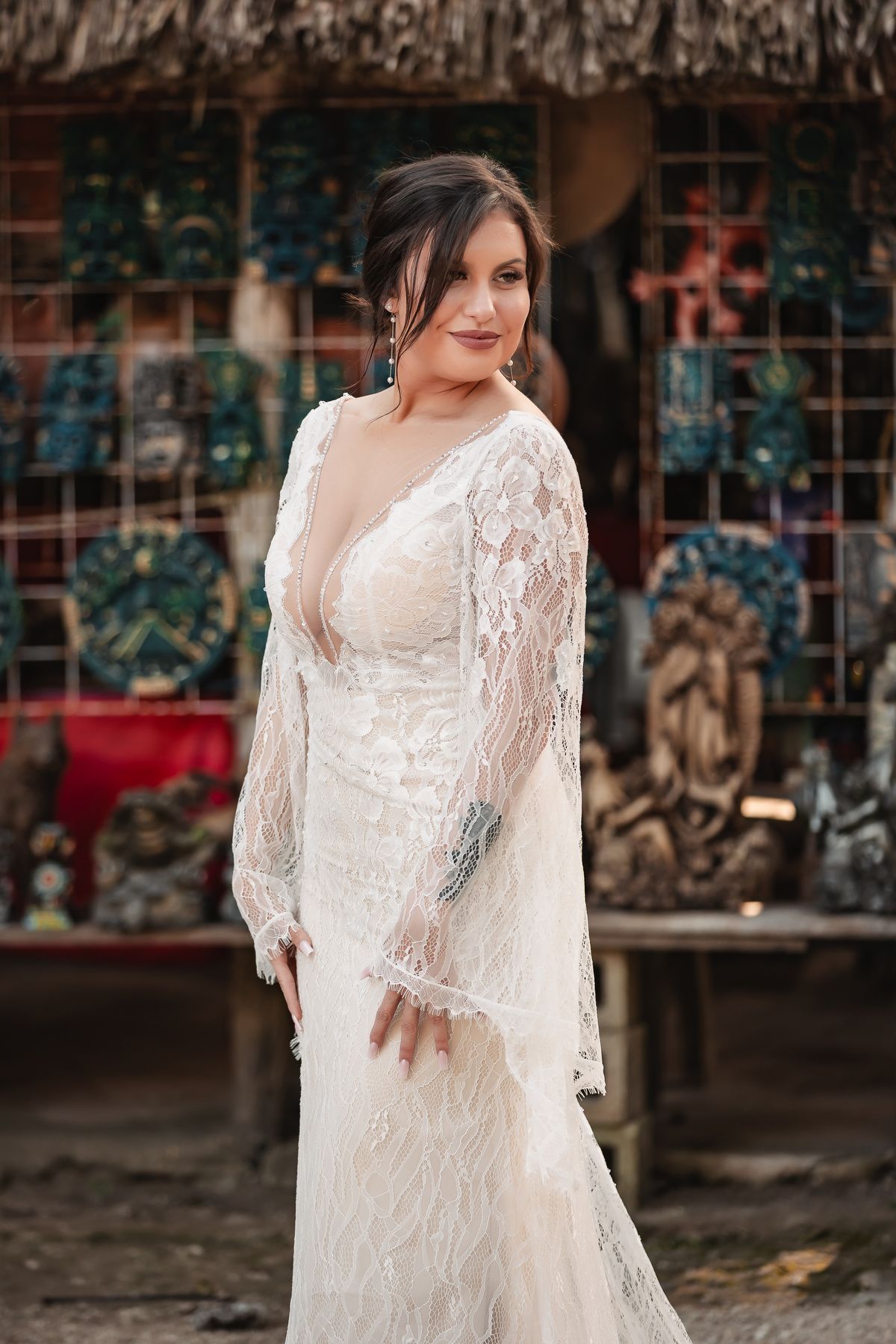 Elegant bride in flowing white lace wedding dress poses outdoors against rustic backdrop with vintage windows.