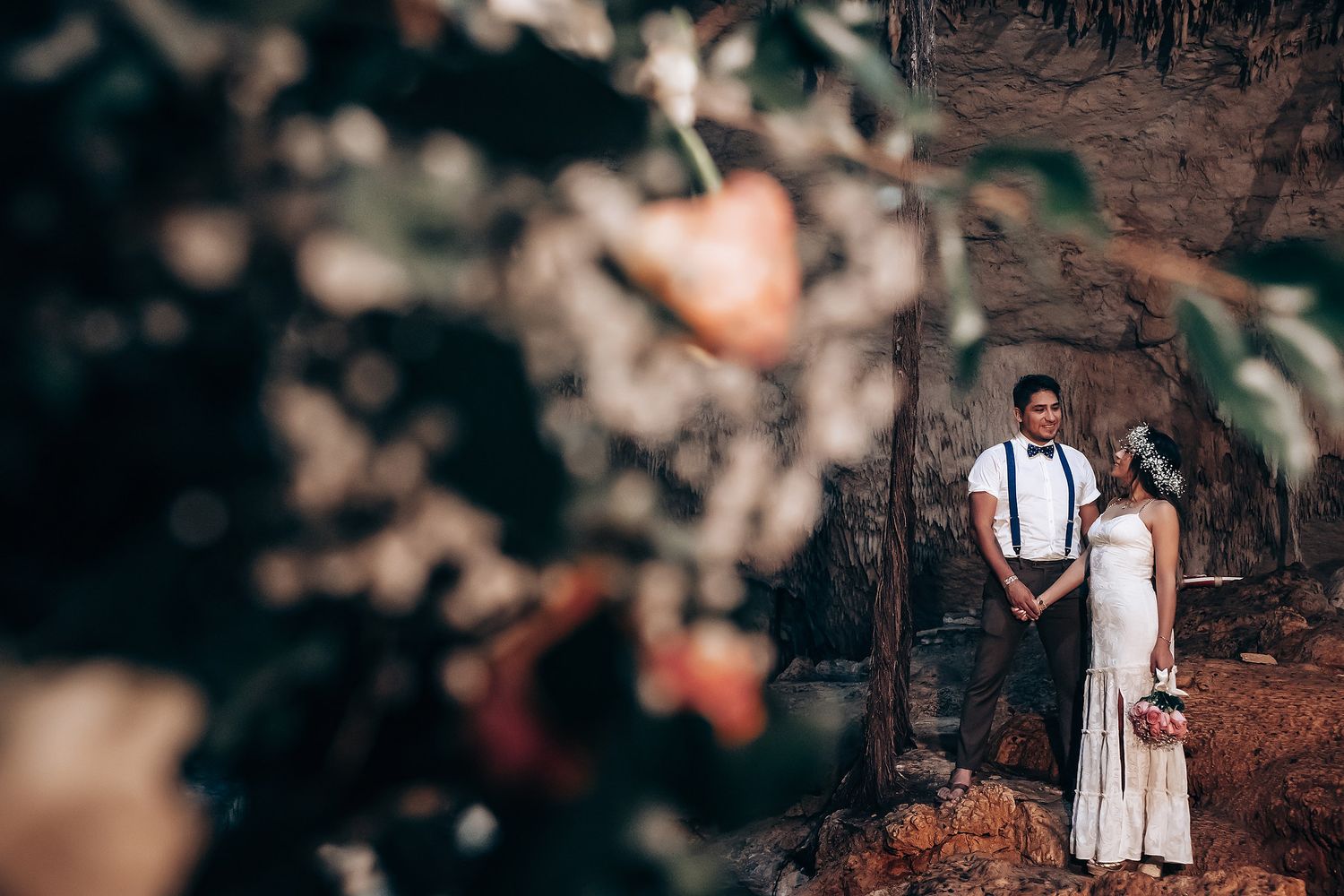 A wedding couple poses against a dramatic cave backdrop with soft floral elements in the foreground.