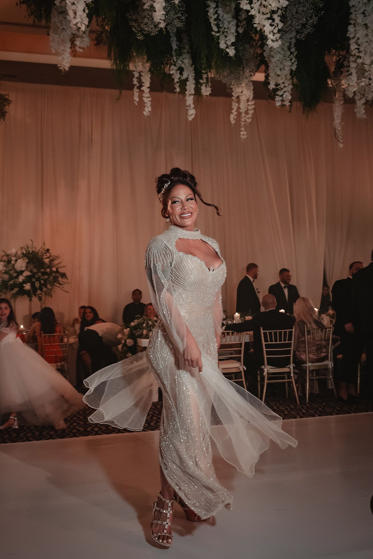 A bride in a white lace dress twirls on the dance floor under hanging floral decorations at a wedding reception.