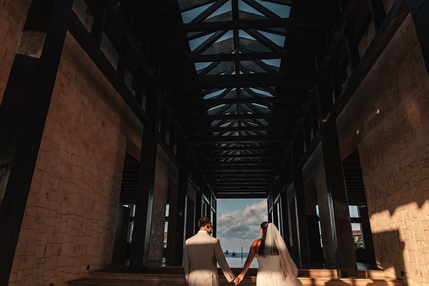 Artistic wedding photo taken through a dark architectural corridor with bright sky visible.