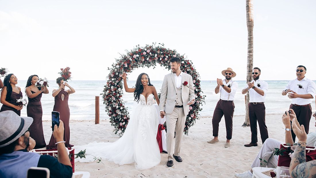Wedding guests gathered under a floral arch on a sunny beach.